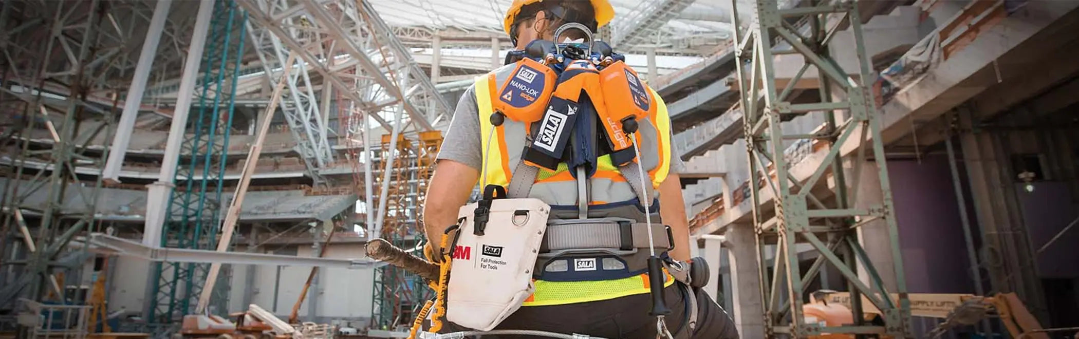 A construction worker is viewed from behind, wearing a high-visibility vest, helmet, and a full-body safety harness at a large indoor construction site. He has several pieces of safety equipment and tool bags attached to his harness, including orange and white DBI-SALA brand accessories.