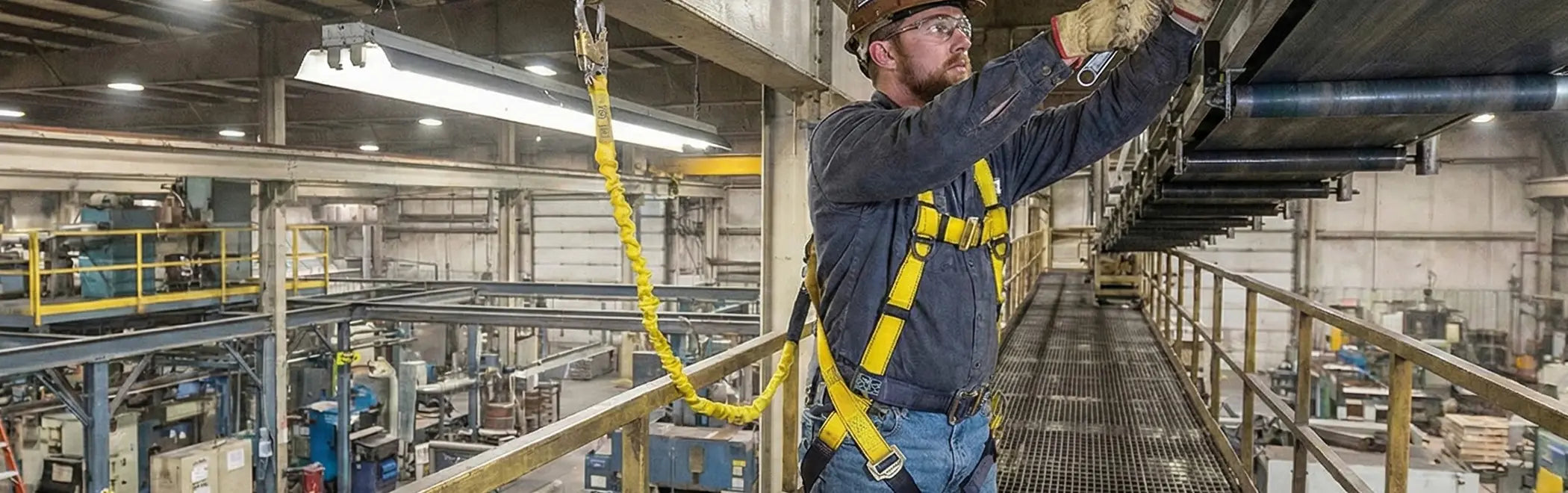A manufacturing worker in a hard hat and safety glasses is performing maintenance on overhead equipment in an industrial indoor facility. He is secured by a yellow full-body safety harness and a matching yellow lanyard attached to an anchor point above a metal grate walkway.