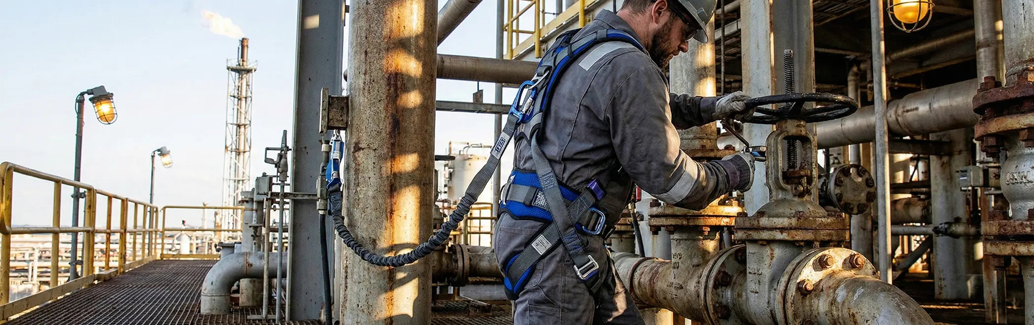 An oil and gas worker in a grey jumpsuit and helmet is operating a large industrial valve on complex piping at a refinery or processing plant. He is wearing a blue and grey full-body safety harness with an attached self-retracting lifeline in an outdoor setting.