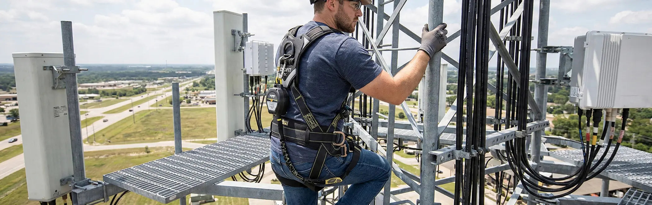 A telecommunications worker, wearing a dark blue shirt and a black full-body harness, is climbing a metal support structure on a cell tower high above a city landscape. He is surrounded by bundled cables and metal grating platforms, likely secured by a personal fall arrest system.