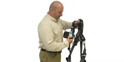 A worker is shown indoors in a light-colored shirt, holding up a full-body safety harness by the back D-ring while inspecting the webbing and tags. This step confirms the harness is free of damage and properly prepared for use before donning.
