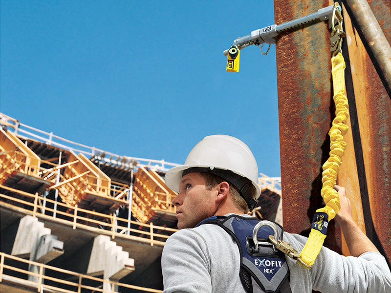 This image shows a worker using a temporary 3M anchorage connector clamped onto a steel beam on a construction site. The connector serves as a direct attachment point for the worker's lanyard to secure them while working at height.