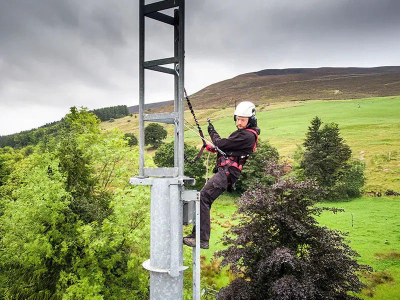 A climber scales a communications tower located on a remote, grassy hill. The worker is securely attached to the structure, showcasing the use of 3M fall protection equipment in climbing applications.