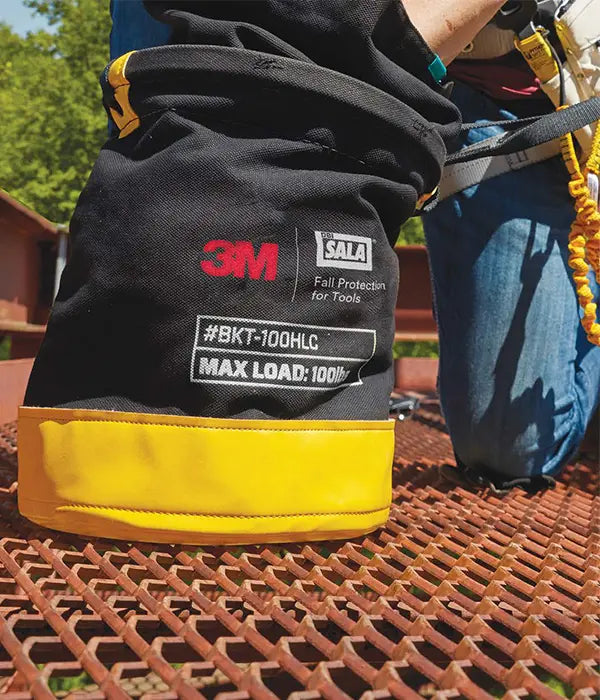 A close-up shot of a black and yellow 3M DBI-SALA tool bucket being used by a worker kneeling on a metal grate surface. The markings on the side indicate the model #BKT-100HLC and a MAX LOAD: 100lb limit.