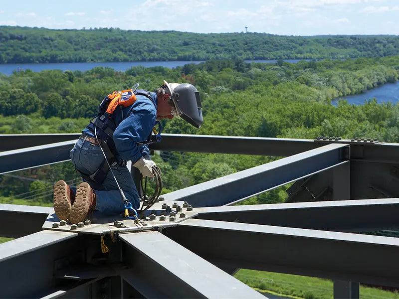 A worker is kneeling on an elevated, open-grid steel platform high above a scenic landscape featuring a river. The worker's gear highlights the necessity of 3M fall protection when performing tasks near edges.