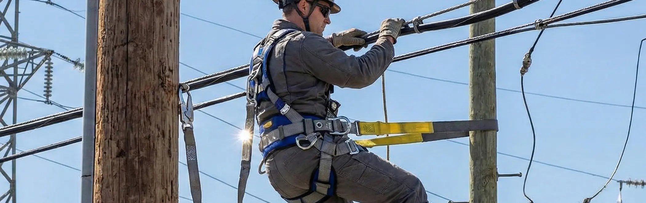 A utility worker wearing a full-body harness and helmet is climbing a wooden utility pole while holding onto electrical cables overhead. He is also secured by a yellow and black positioning lanyard around the pole to safely maintain his elevated position.