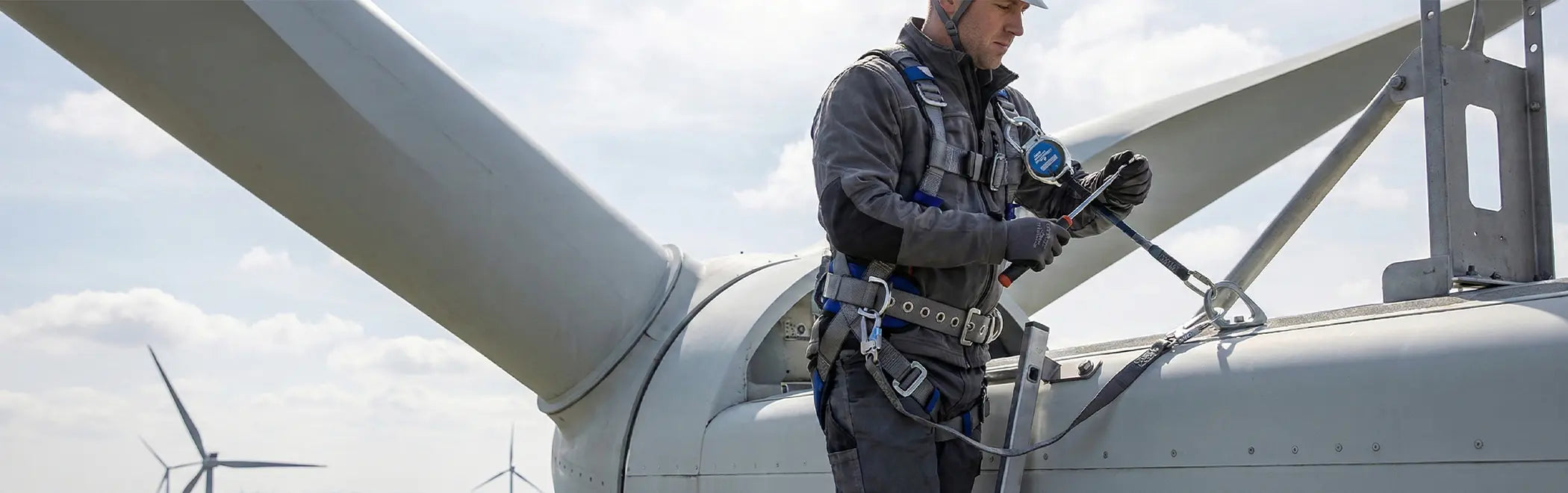 A technician is standing on a large wind turbine blade, adjusting equipment with a tool under a bright, cloudy sky. He is wearing a grey and black full-body harness and a helmet, secured to an anchor point on the turbine structure.